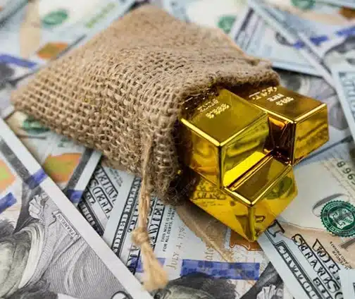 A burlap sack spilling shiny gold bars onto a scattered pile of U.S. hundred dollar bills.