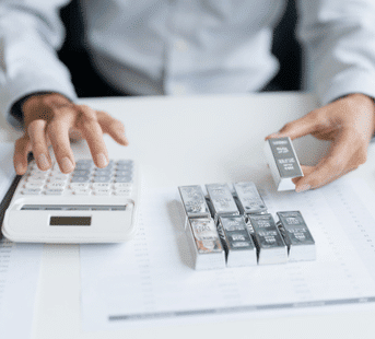 A person uses a calculator with one hand and holds a silver bar with the other. Several silver bars are arranged on a desk along with documents.