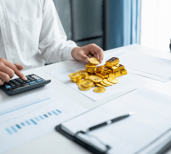 A person in a white shirt uses a calculator at a desk with financial charts, gold coins, and gold bars stacked on top of some documents.