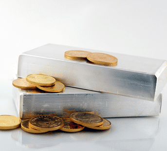 Two large silver bars are stacked on top of each other, with several gold coins scattered on and around them on a reflective white surface.