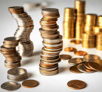 Stacks and piles of assorted coins, including silver and gold colors, are arranged on a white surface. Some stacks are neat, while others are leaning or slightly uneven. Loose coins are scattered around.