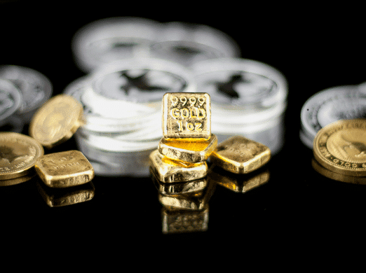 Small gold bars stacked in front of blurred silver coins, all resting on a reflective black surface. The front gold bar is inscribed with "9999 GOLD 1 OZ.