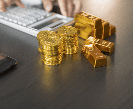 Stacks of gold coins and gold bars are arranged on a dark desk next to a tablet, while a person types on a calculator in the background.