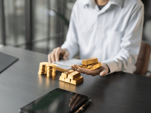 A person in a white shirt sits at a desk, holding gold bars in one hand with more gold bars and stacks of gold coins on the table; a tablet lies in the foreground.