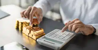 A person stacks gold bars and coins on a desk while using a calculator, symbolizing financial planning or investment in precious metals.