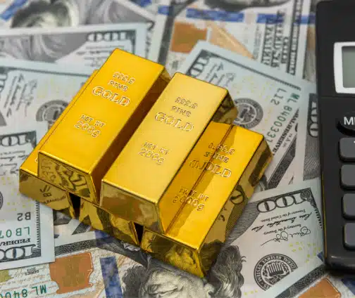 A burlap sack spilling shiny gold bars onto a scattered pile of U.S. hundred dollar bills.